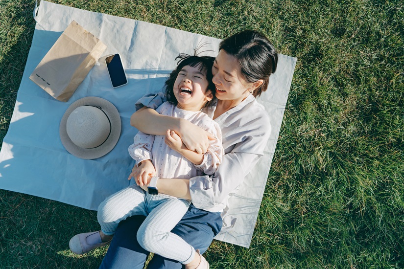 Affectionate young Asian mother embracing little daughter in arms, lying down on the grassy field, having fun and smiling joyfully, enjoying together on a sunny day. Family love and bonding time. Enjoying the nature. Outdoor fun concept