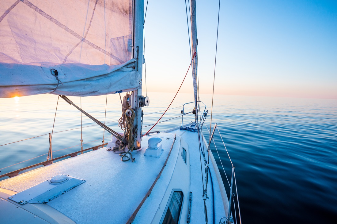 The calm water. White sloop rigged yacht sailing at sunset. A view from the deck to the bow, mast and the sails. Baltic Sea, Latvia
